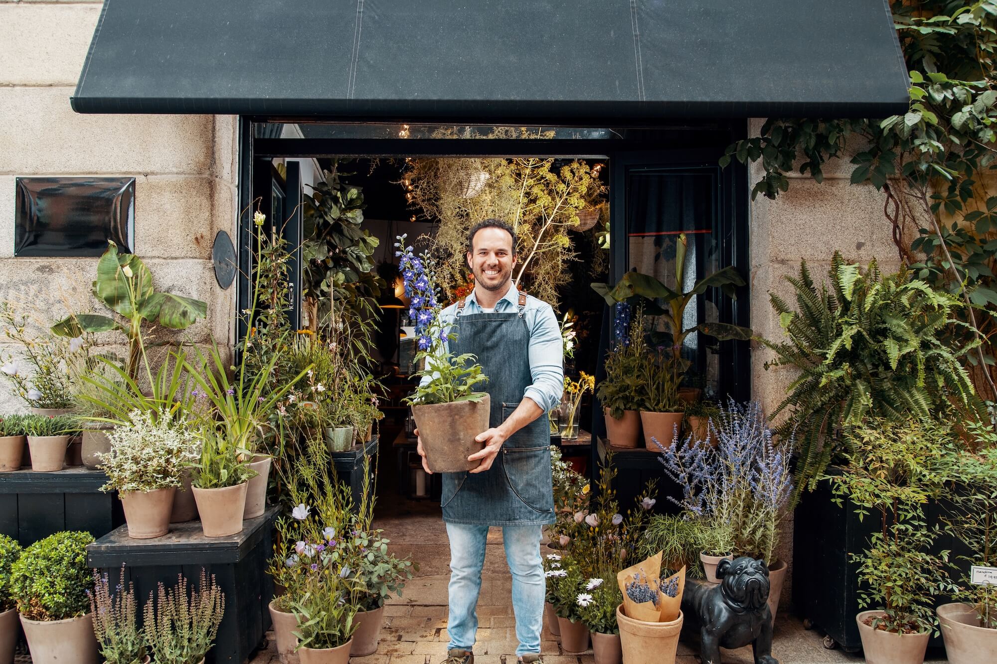 Smiling shop owner standing outside a lush, plant-filled storefront holding a potted flower, symbolising Supr Bueno’s mission to empower local businesses with beautifully crafted, conversion-focused Shopify websites.